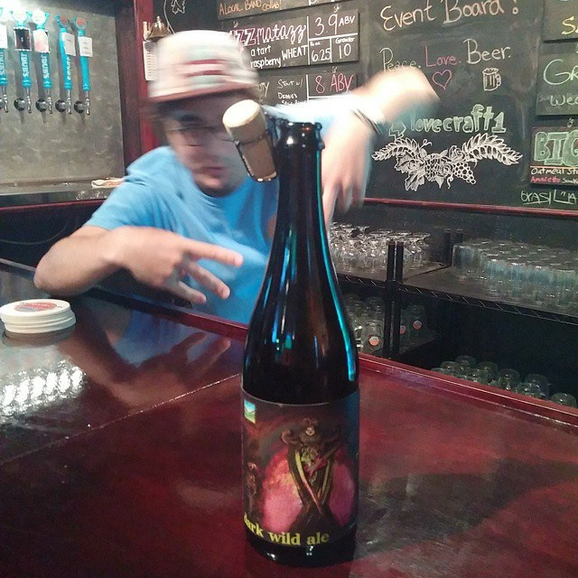 A bottle of wild fermented ale from an Indiana brewery sits on a bar; a bartender in a blue shirt and hat stands behind the bar.