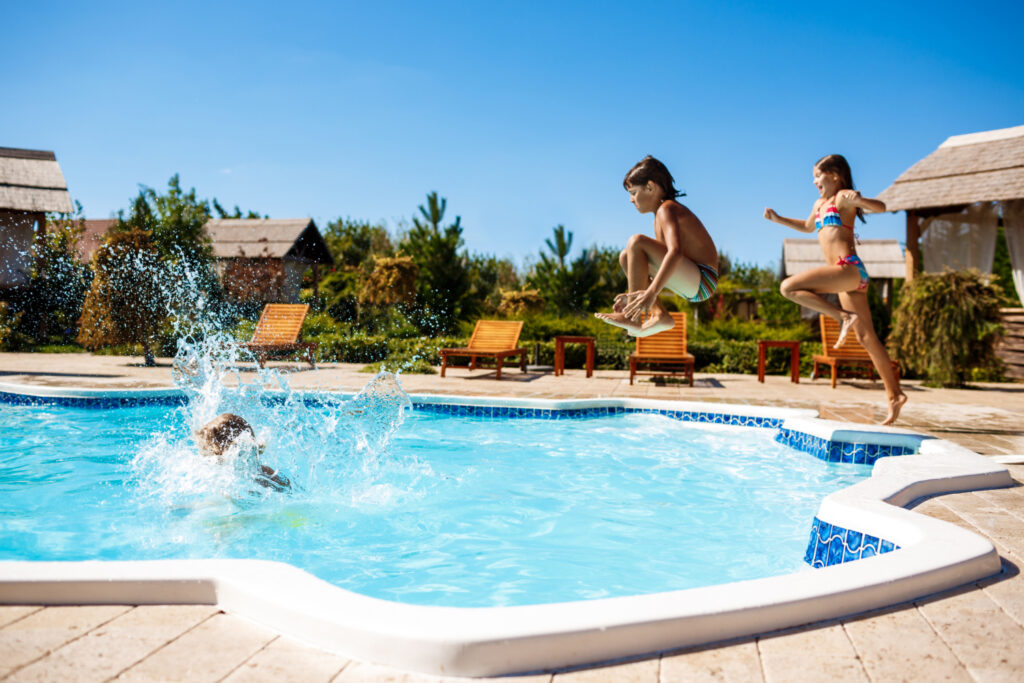 three young kids jump into an outdoor hotel pool, causing water to splash into the air.