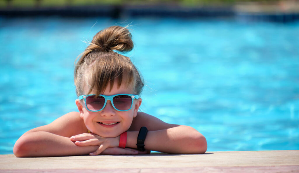 A little girl wearing sunglasses and her hair tied up into a high bun smiles as hangs on to the side of the pool wall.