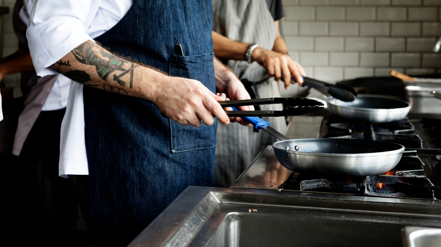 A restaurant chef stands at a stovetop, holding a sauce pan over open flame and tongs. He's wearing a dark demin blue apron and white shirt. Forearm tattoos are visible from under his chef's jacket sleeve.