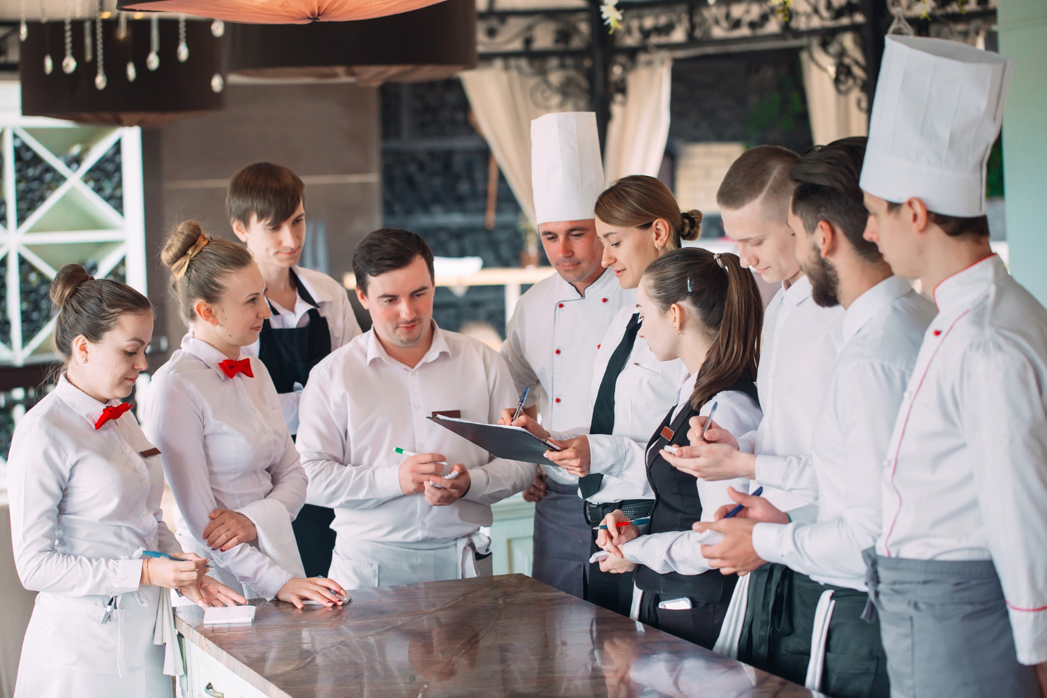A femal restaurant manager holding a clipboard is surrounded by her restaurant employees, holding a team meeting. The employees are dressed in black and white, some wearing chef coats and hats, others wearing white aprons.