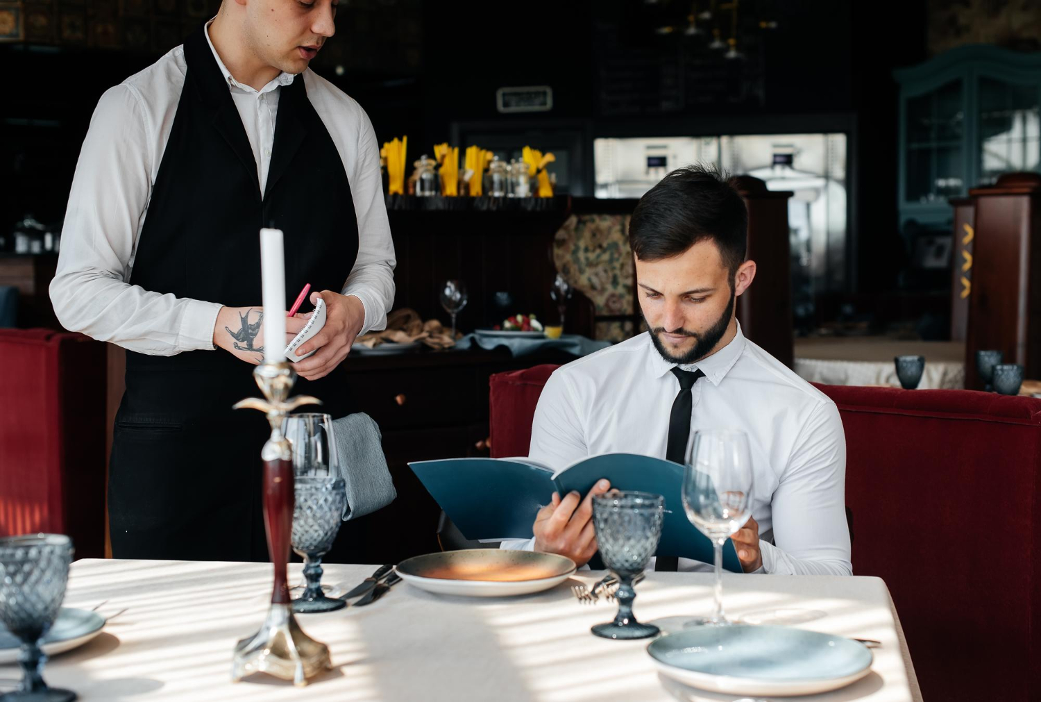 A male waiter stands next to man seated at a restaurant table. The man sitting down is wearing a white shirt and black tie and is reading a menu while the waiter waits to take his order.