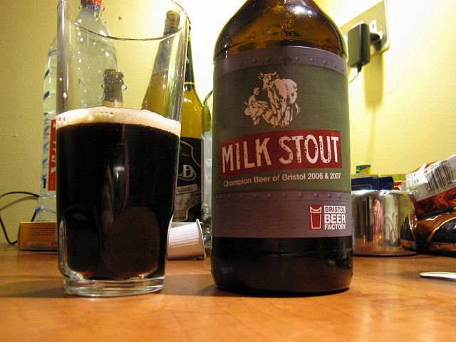 Half a pint of dark milk stout beer in a glass sits next to a beer bottle with a dark green label with the words "milk stout" across the front.