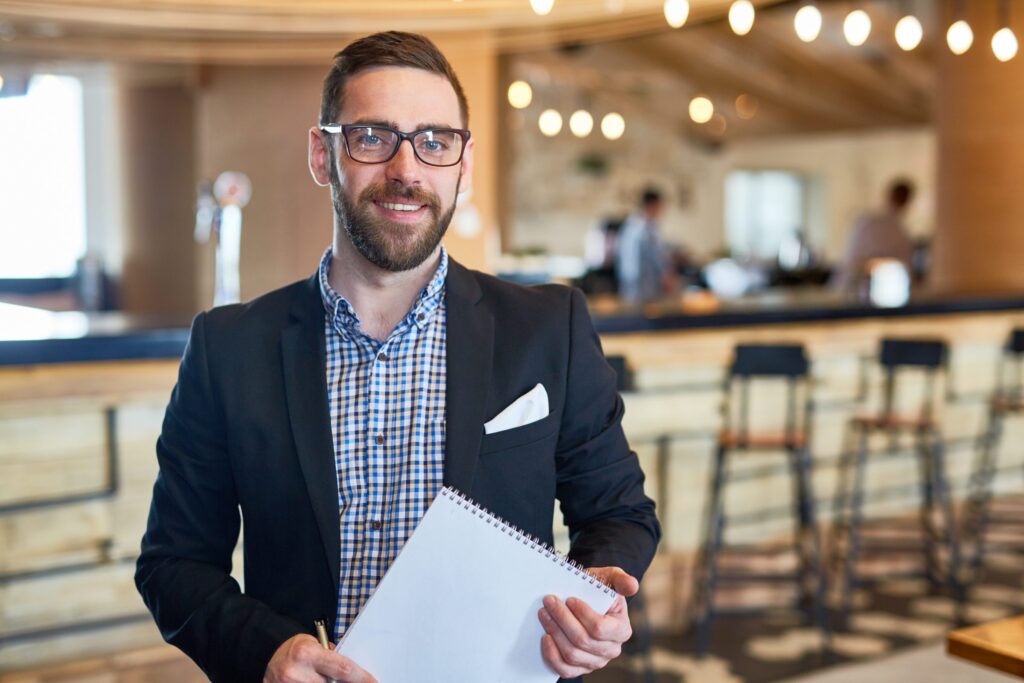 A man standing with a paper pad in a restaurant with a bar in the background.