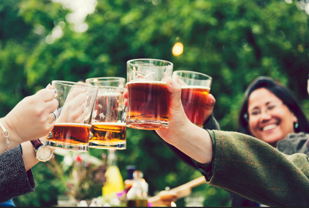 A close-up of pint glasses filled with beer clinking together in a toast outside in a beer garden. A woman participating in the cheers smiles. 