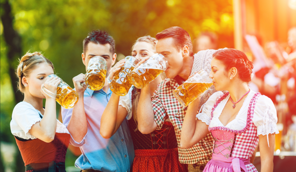 Men and women dressed for Octoberfest drink beer from large glass steins in a friendly outdoor beer garden. 