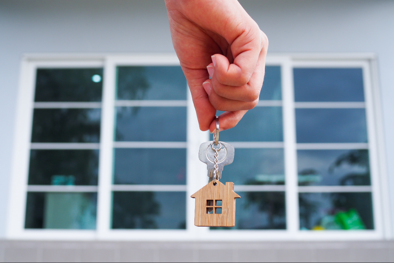 In front of large house windows, a manicured hand dangles keys with a small house token on the keyring. 