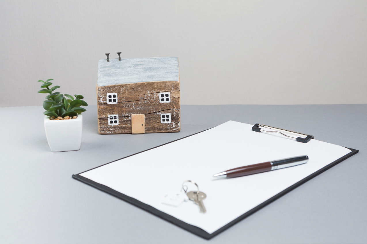 A miniature model of a home sits on a gray desk next to a small potted plant, a clipboard with a pen, and the house keys.