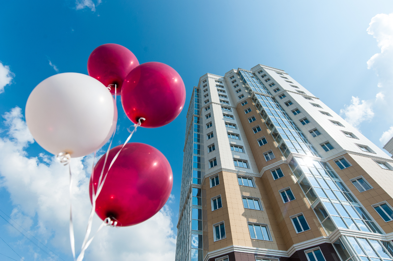 A photo of a tall apartment building stretching into the blue sky. Red and white balloons floating nearby allude to a new lease celebration. 