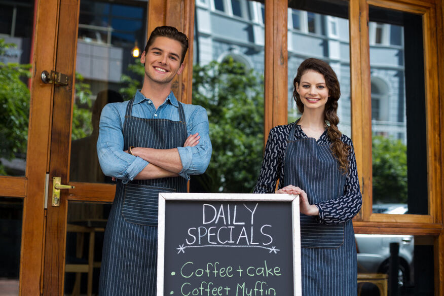 A smiling man and woman stand outside their establishment next to a sandwich board that lists the daily specials, which is a restaurant industry trend. 