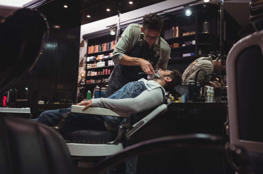 A barber carefully shaves a client’s face using a straight razor in a dark, comforting salon in front of a shelf of hair products. 