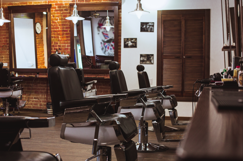 A line of empty salon chairs are placed in front of mirrors in a relaxing, earthy salon with an exposed brick accent wall.