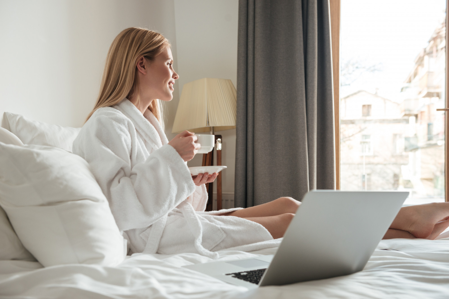 A woman in a hotel robe, sitting on a bed enjoying a cup of morning coffee while looking out at the view outside of her hotel window.