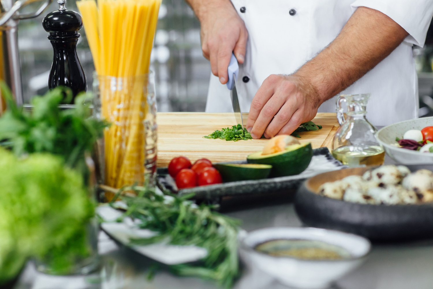A chef in the kitchen of a restaurant chopping herbs on a cutting board, surrounded by pasta, greens and other fresh food.