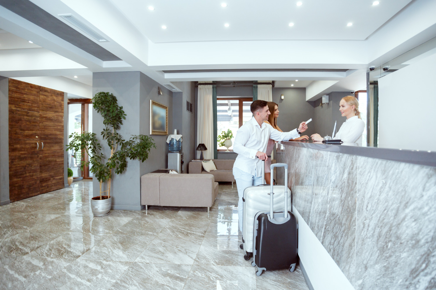 A couple stands at the front desk in a hotel lobby with their suitcases, talking with the front desk clerk and checking in to their hotel room.