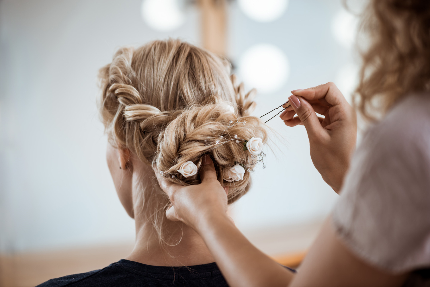 A hairdresser works on a bride's hair, tied up into a braided bun and pinned in place with rose-decorated pins and beads.