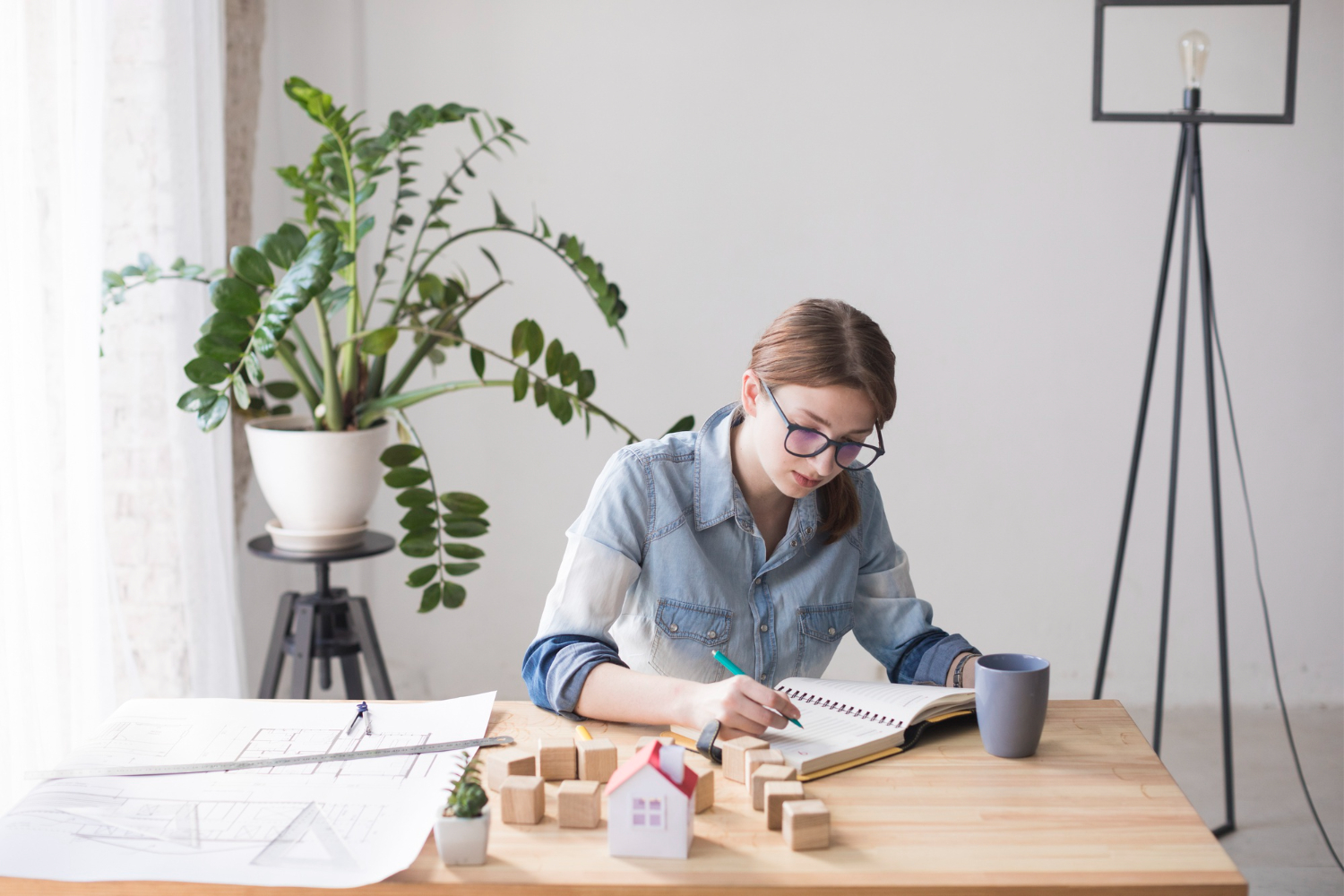 A co-op building manager sits at a table, writing in a notebook, with a small model house, building blocks, and apartment plans spread out on the table in front of her.