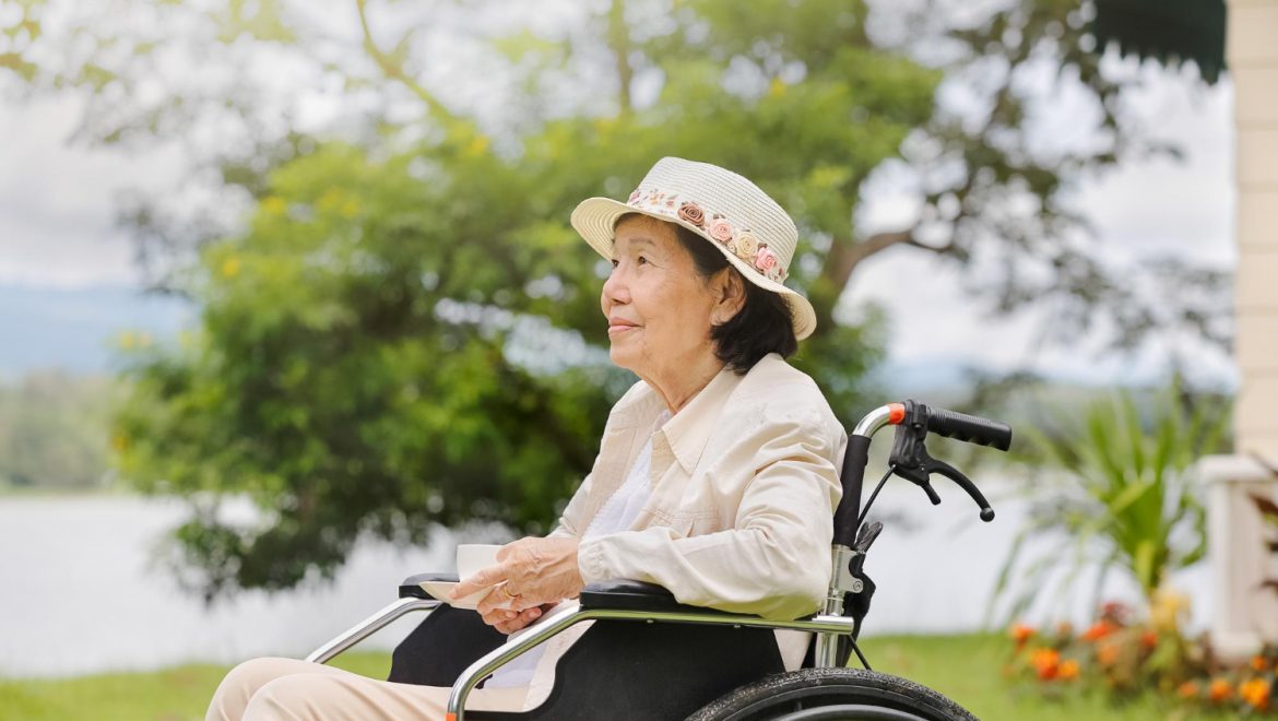An elderly woman sits outdoors in her wheelchair, admiring the lush green space and foliage around her.