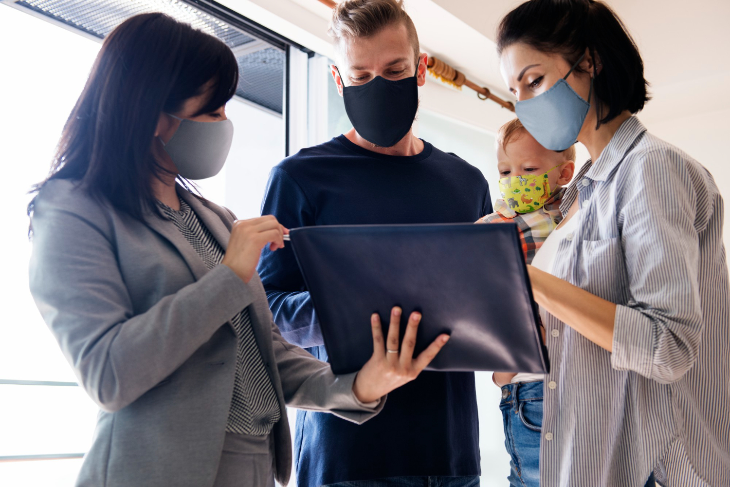 A realtor, husband, wife, and young baby all wearing face masks, gather around a large tablet screen, watching a virtual guided apartment tour.