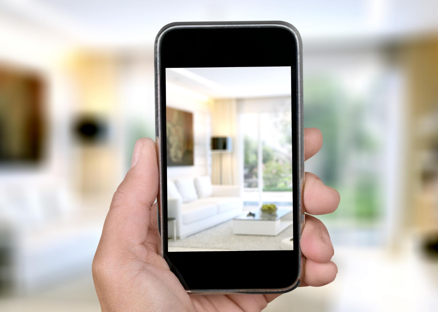 A man holds a smart phone with an out-of-focus view of of a living room on the screen, giving a virtually guided apartment tour,