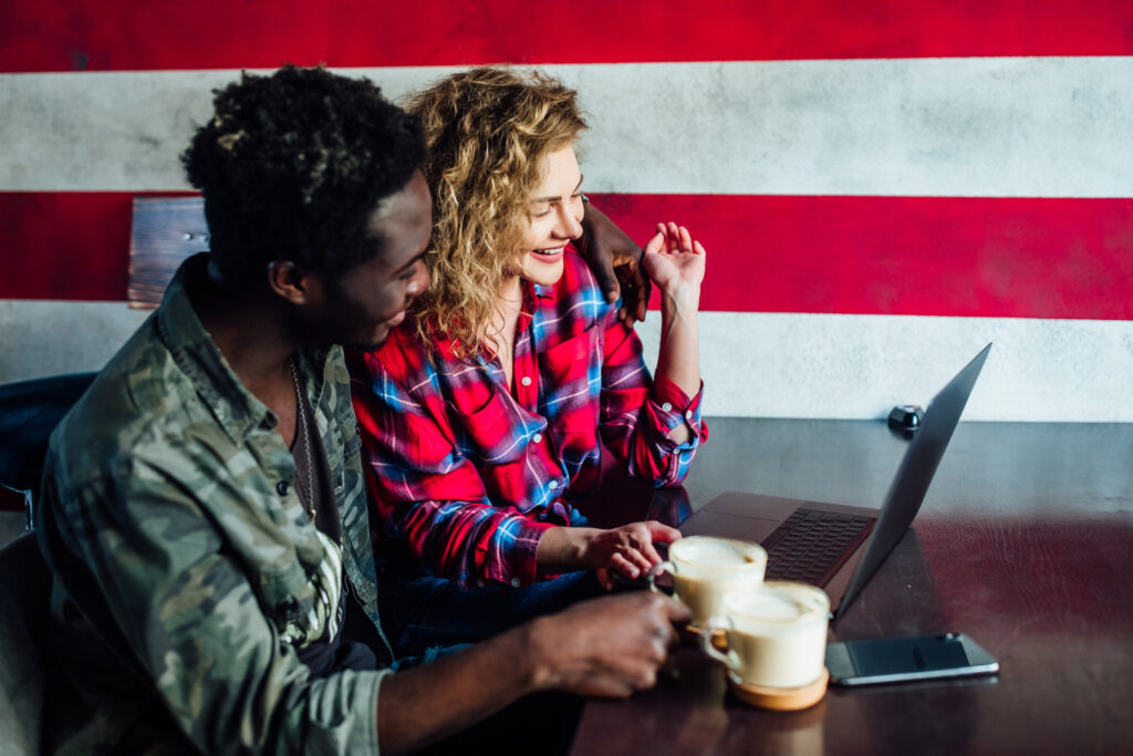 A smiling young man and woman sit in a coffee shop, looking at a laptop on the table, watching a virtual apartment tour as out-of-state renters.