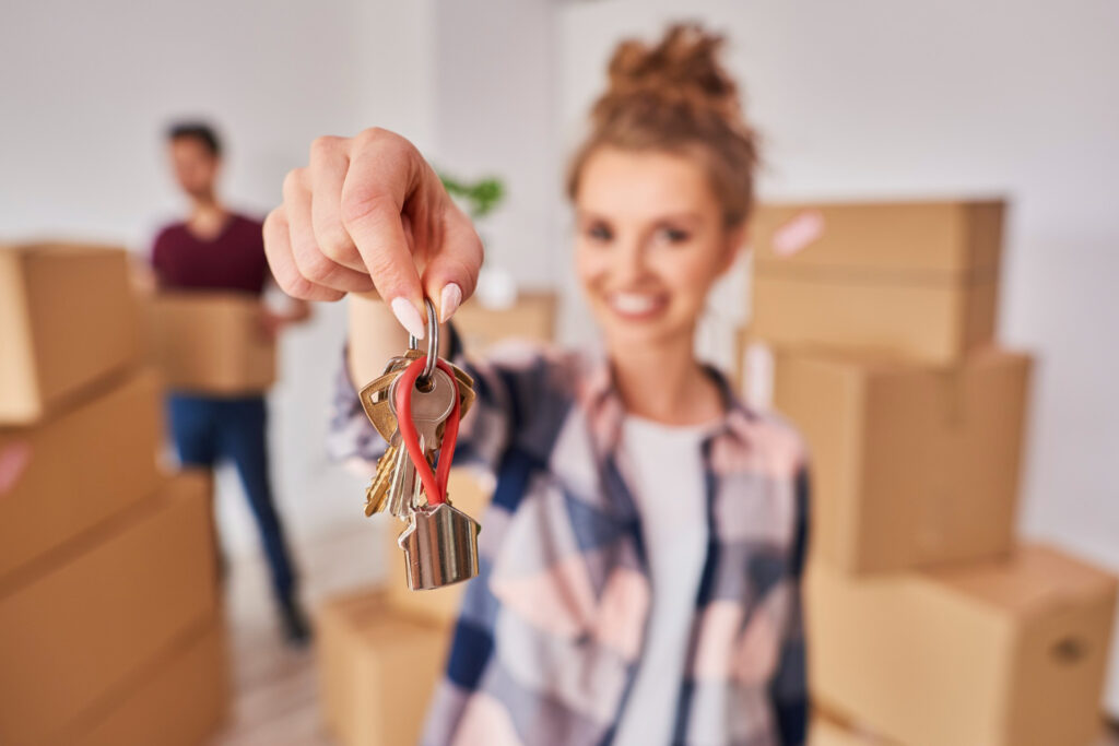 A woman holds a set of apartment keys towards the camera, surrounded by moving boxes in her new apartment.