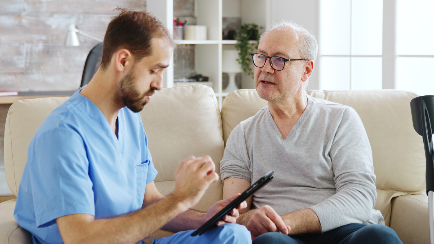 An older adult man wearing a grey shirt sits next to an assisted living nurse wearing blue scrubs. The nurse is holding and looking at a tablet.