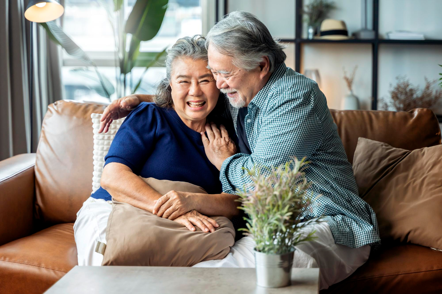 An older couple sits on a brown leather couch, hugging and smiling at the camera.