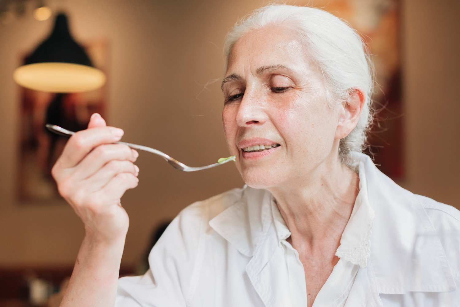An older lady with white hair, wearing a plain white shirt, holds a fork with a single bite of food up to her mouth.