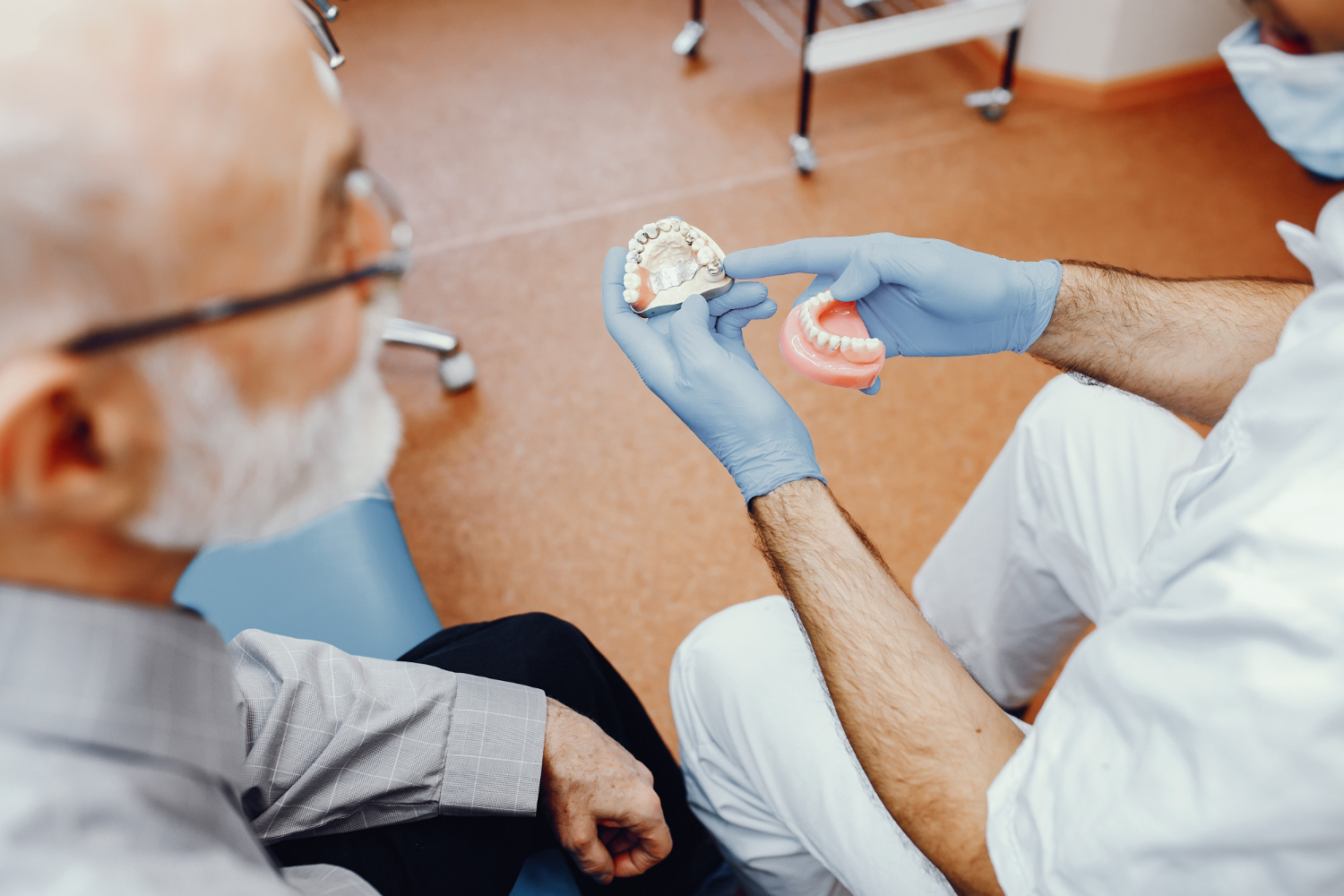 An older man sits with his dentist. The detist is holding a set of dentures and blue medical gloves, pointing at the detures and speaking to the older man.