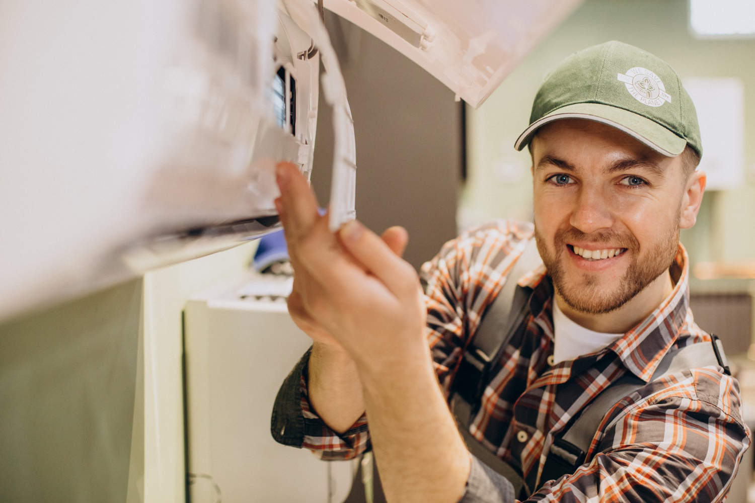 An HVAC technician works on an air conditioning using. He's wearing a brown, white, and orange plaid long-sleeve shirt and an olive green hat, and is smiling at the camera.