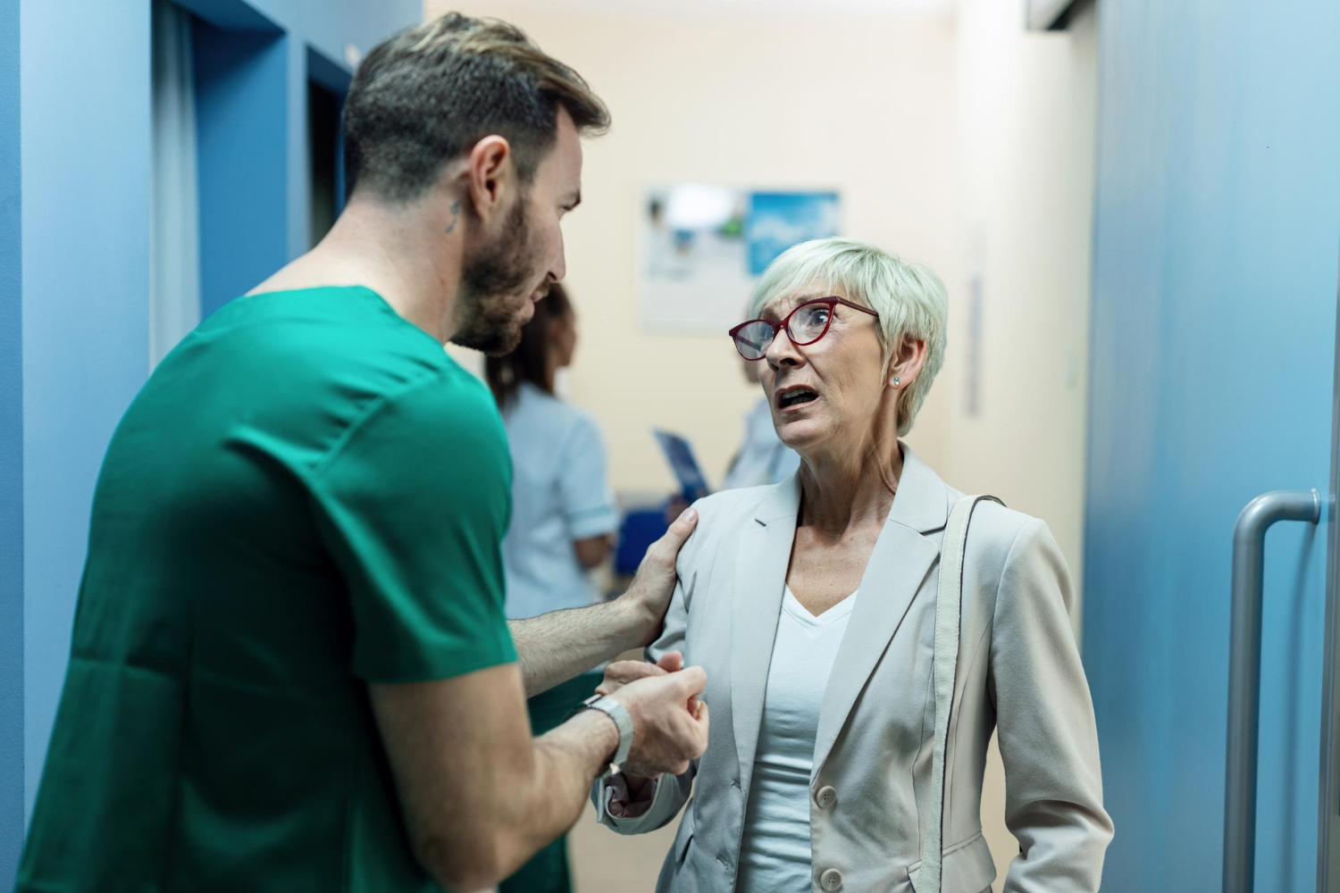 A male medical professional wearing a green scrub top speaks to an older, confused looking woman. He is holding her hand, and has another hand on her shoulder.