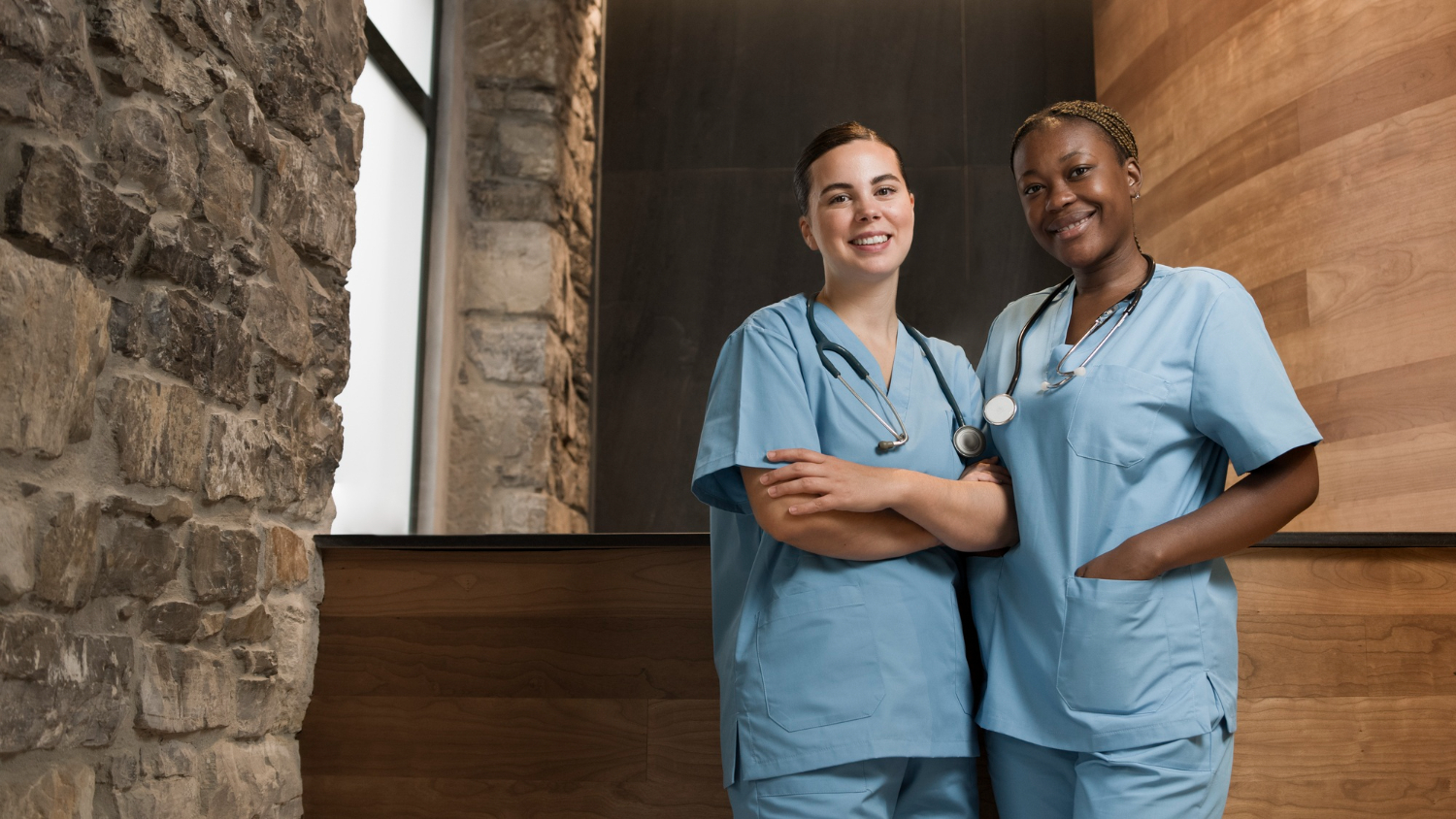 Two female nurses wearing blue srubs stand in a stone and wood-panneled room. One nurse is white with brown hair, and stands with her arms casually crossed, smiling. The second nurse is African American with brown hair. She stands with one hand in the pocket of her scrib top, also smiling at the camera.