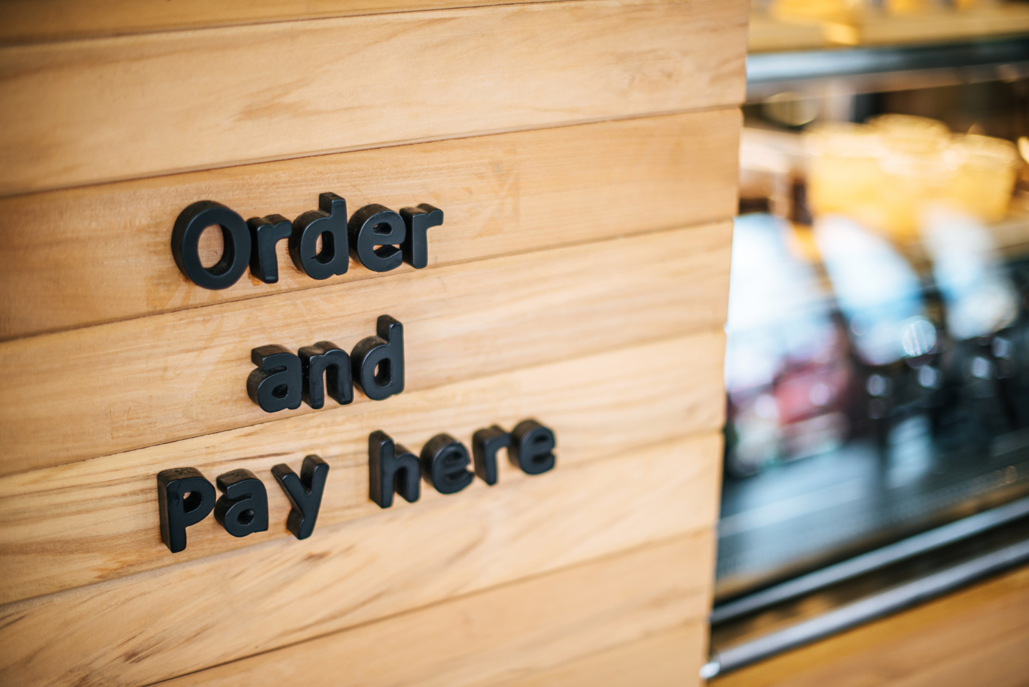 The words "order and pay here" are mounted on a wood wall at a fast casual dining restaurant. A display case full of bakery items is visible, out of focus, in the background.