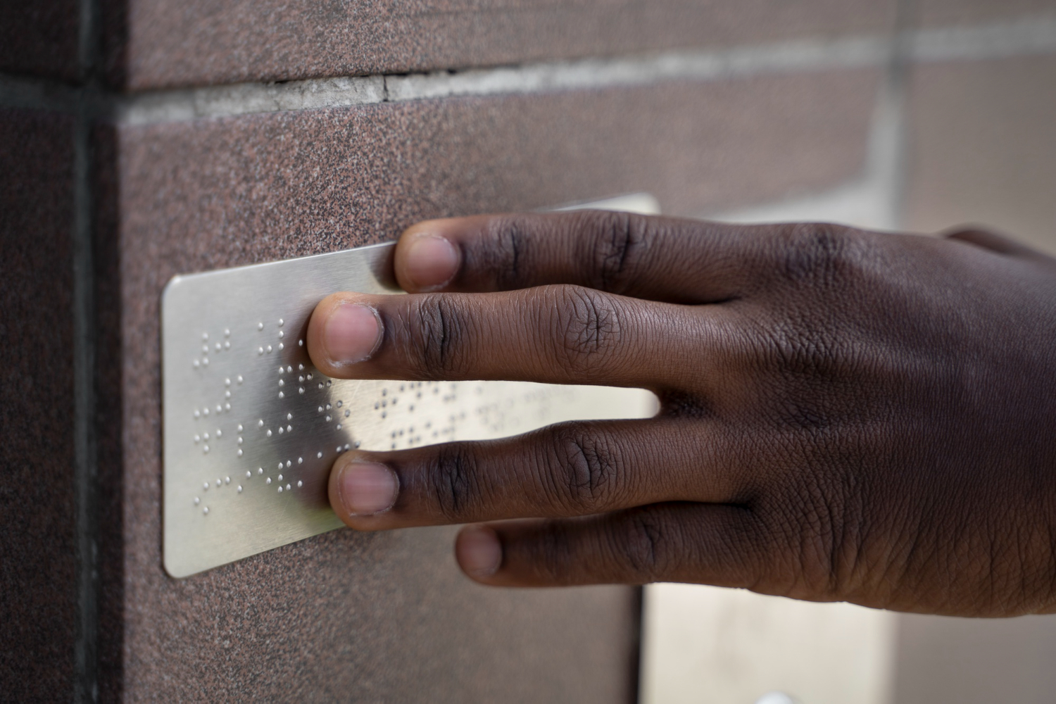 An African American man's hand touches a sign with Braille markings on a brick wall.