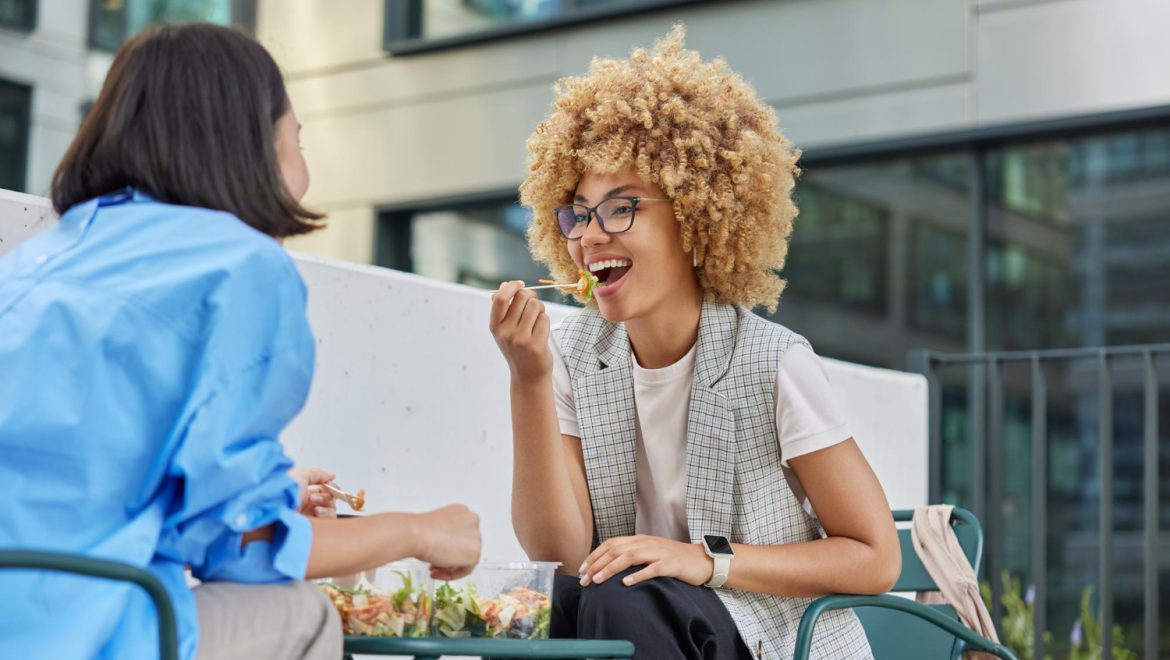 Two women sit outside at an outdoor cafe, engaged in conversation while eating salads.
