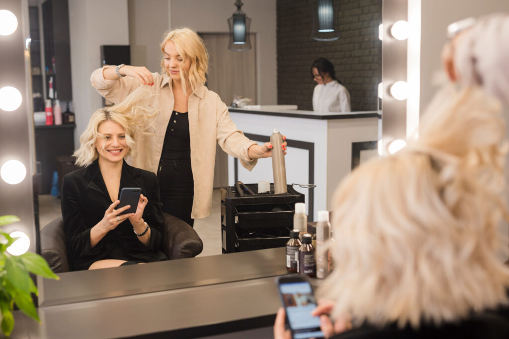 A woman sits in a salon chair, smiling into the mirror as a hair stylist prepares to spray and style her hair. 