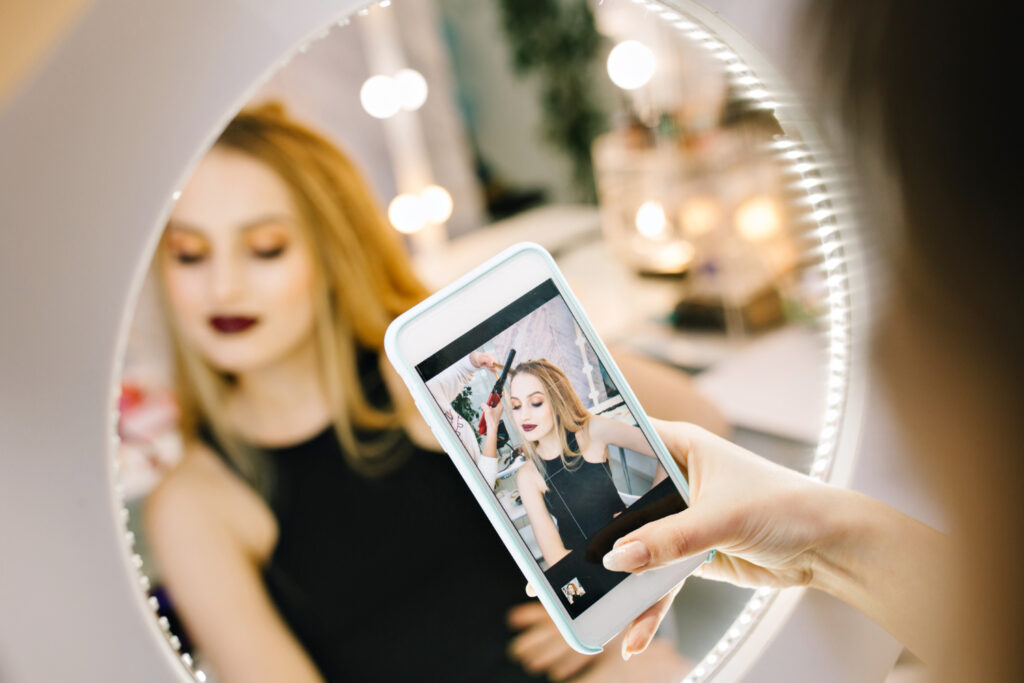 A woman’s hand holds a phone camera up to a mirror. The woman’s reflection, which shows her getting her hair styled, is seen on the phone’s screen. 