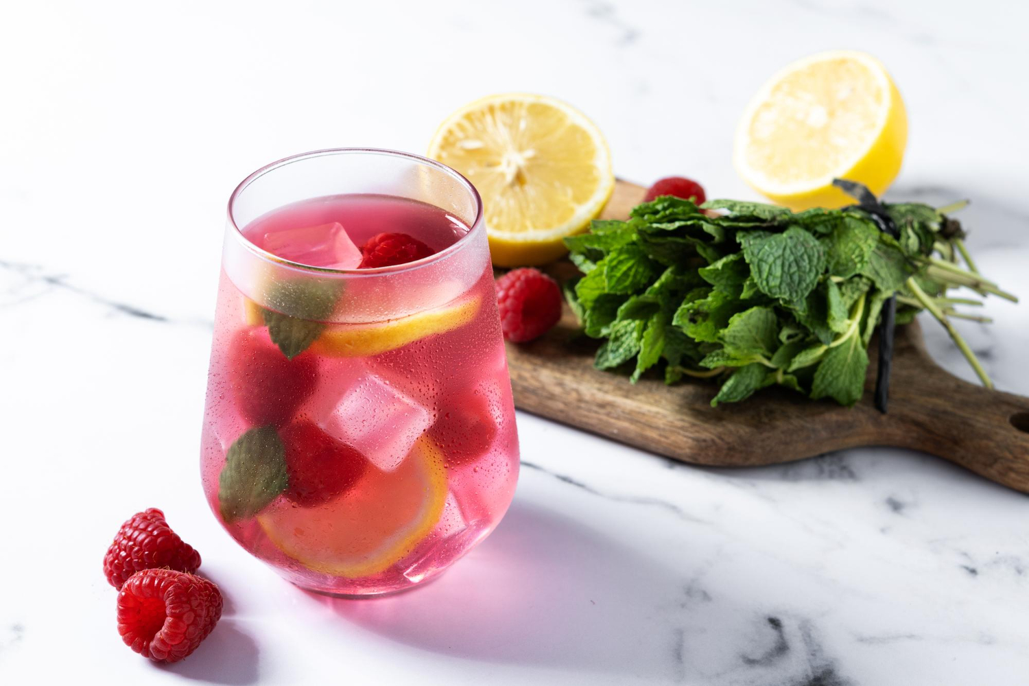 A glass of pink colored hard seltzer garnished with lemon, raspberries, and mint leaves sits next to a small serving board filled with a bunch of mint leaves, a lemon sliced in half, and fresh raspberries.