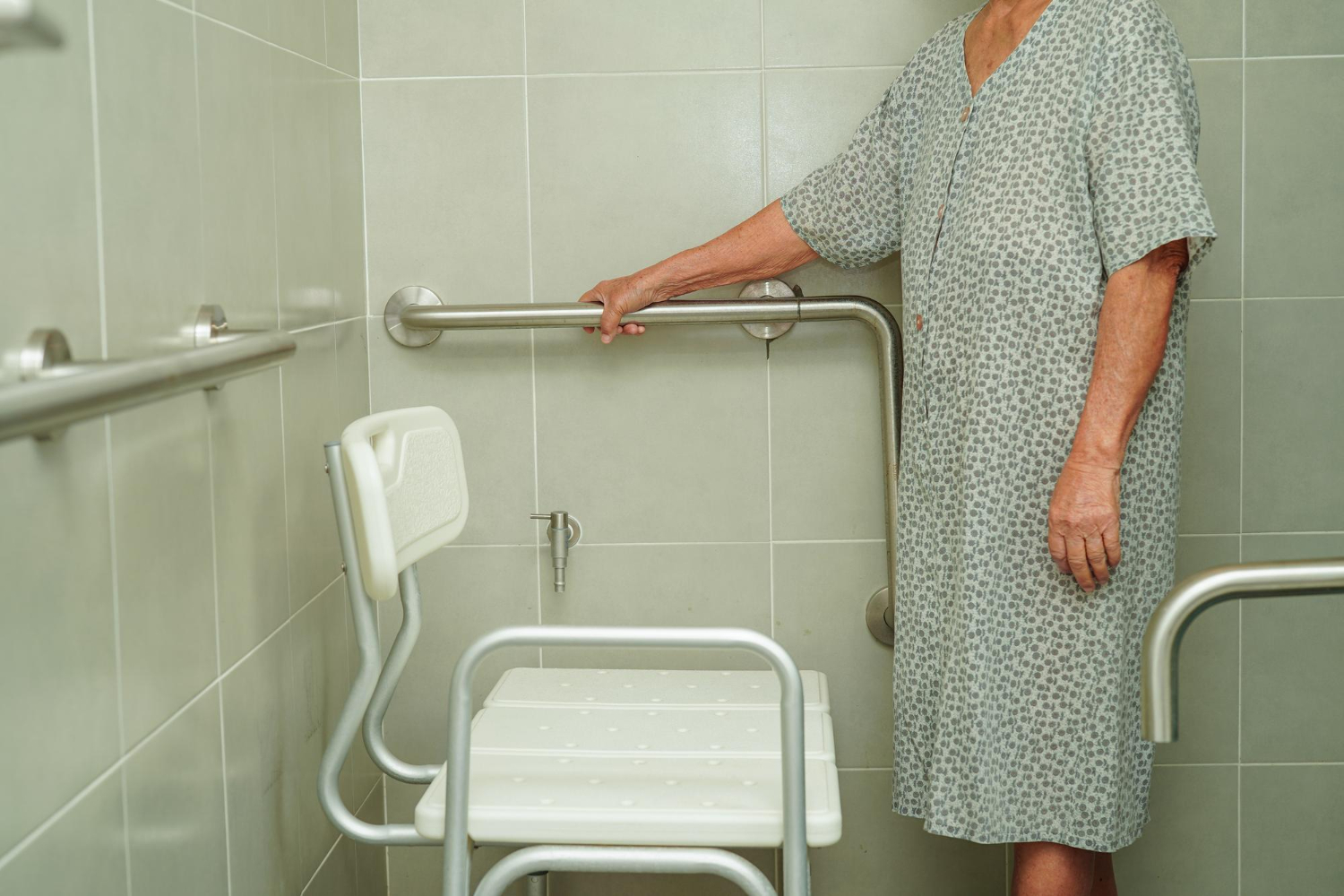 An elderly woman in a nightgown stands inside a large walk-in shower. She's holding on to a wall-mounted metal grab bar, and standing in front of white shower chair.