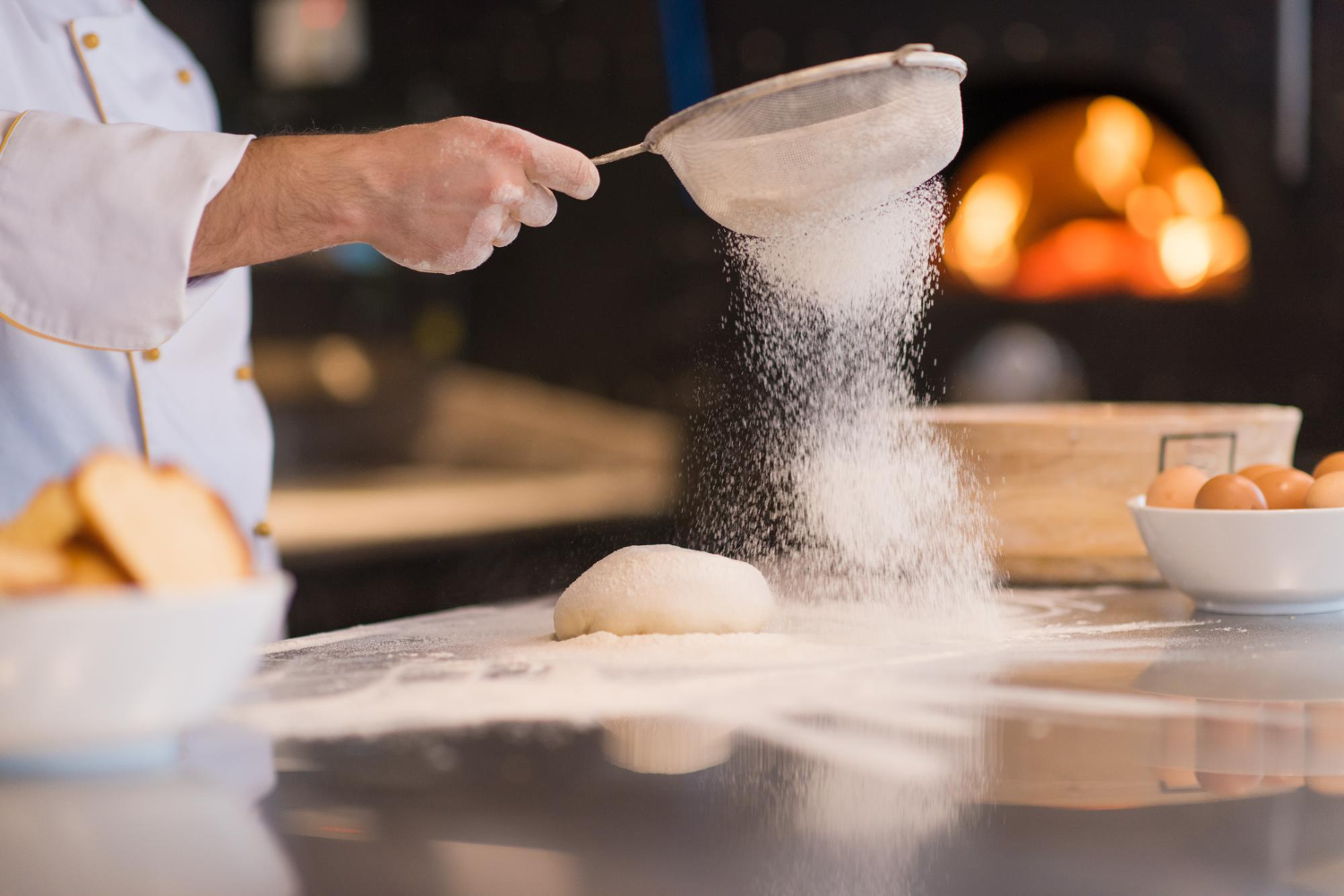 A chef sprinkles flour from a mesh shifter over a ball of bread dough on a stainless steel countertop.