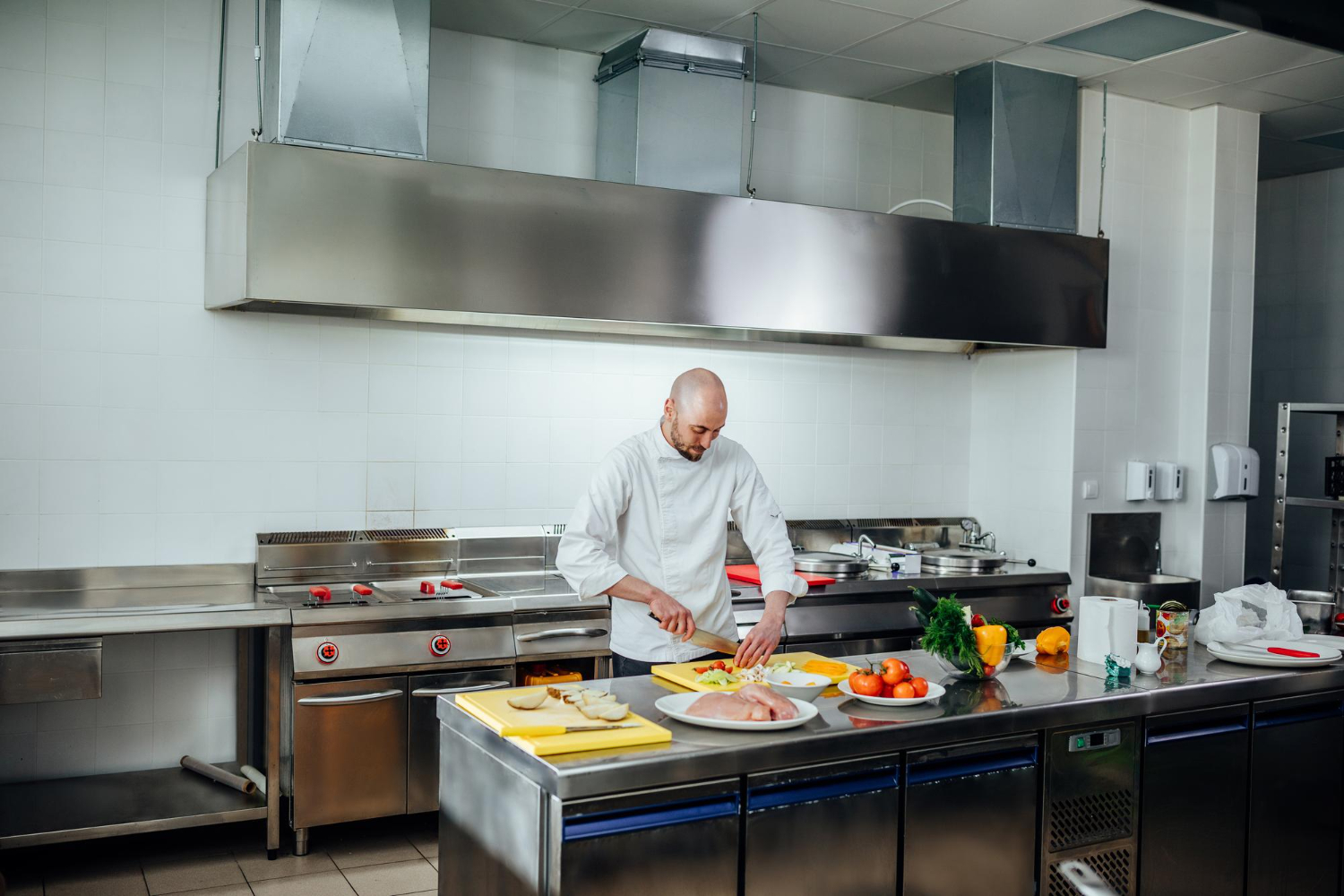 A chef works at a prep table in a commercial kitchen, slicing vegetables and preparing food on cutting boards. Behind the chef is a row of stainless steel fryers and industrial exhaust fans.