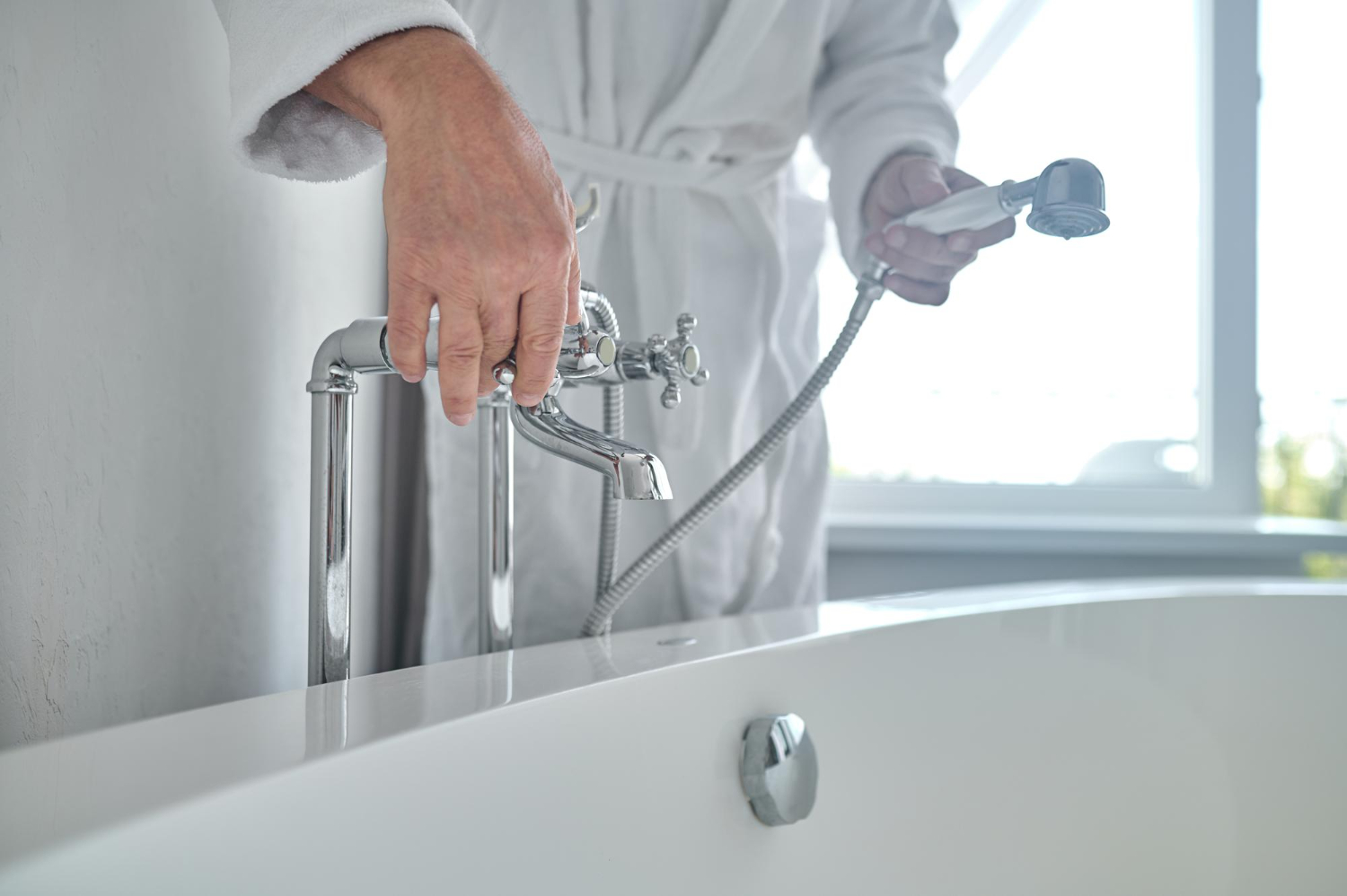 A man wearing a white bathrobe turns the handles of a bathtub faucet. He holds a detectable showerhead in his other hand.