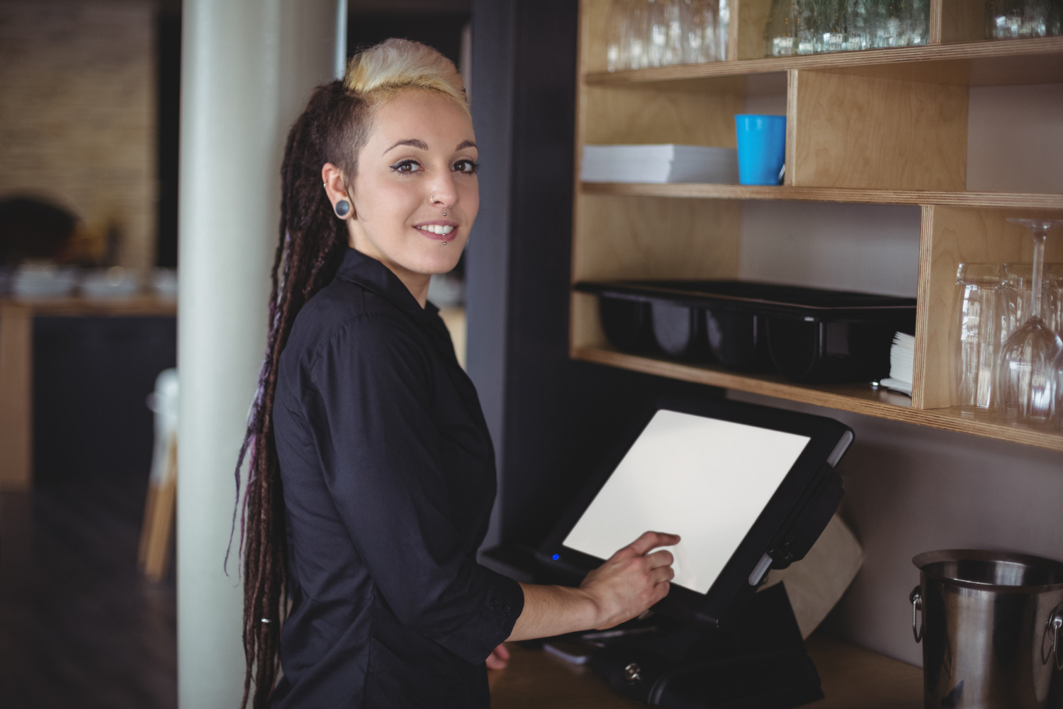 A waitress with long black and blond hair stands at a restaurant POS system, inputting a customer order. She's wearing all black, and is looking over her shoulder at the camera.