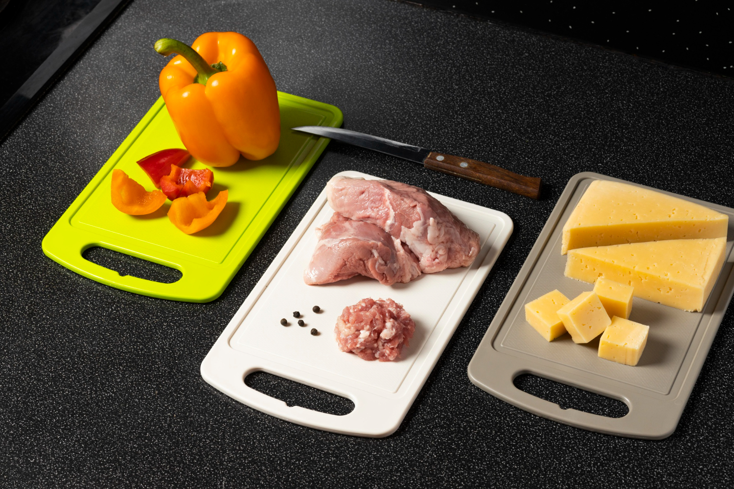 Sliced orange bell peppers, raw chicken, and cubed cheese each on a different cutting board to prevent cross-contamination. The cutting boards are green, white, and brown, and sit on a black countertop.