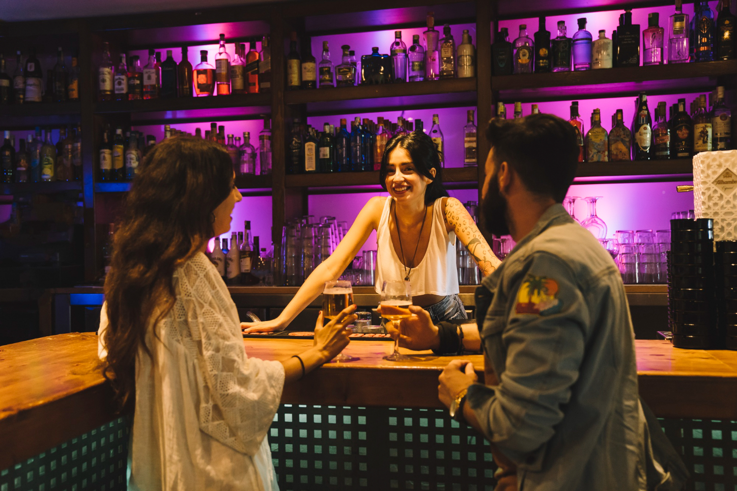 A woman bartender stands behind a bar, chatting with two patrons on the other side of the bar.