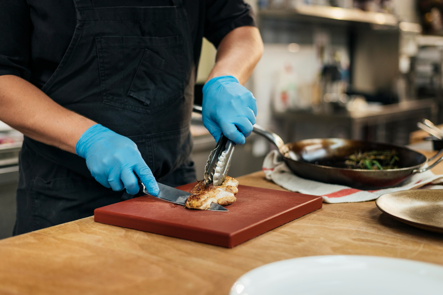 A male chef wears blue food-safe gloves and cuts a piece of cooked chicken on a cutting board in a restaurant kitchen.