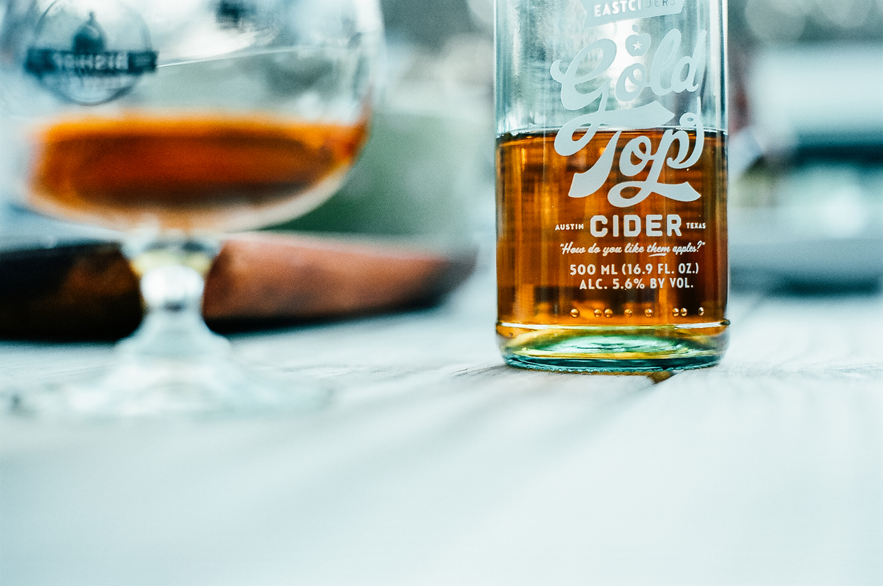 A half empty bottle of hard cider sits on a table. The brewery logo and alcohol content are printed on the glass bottle in white text.