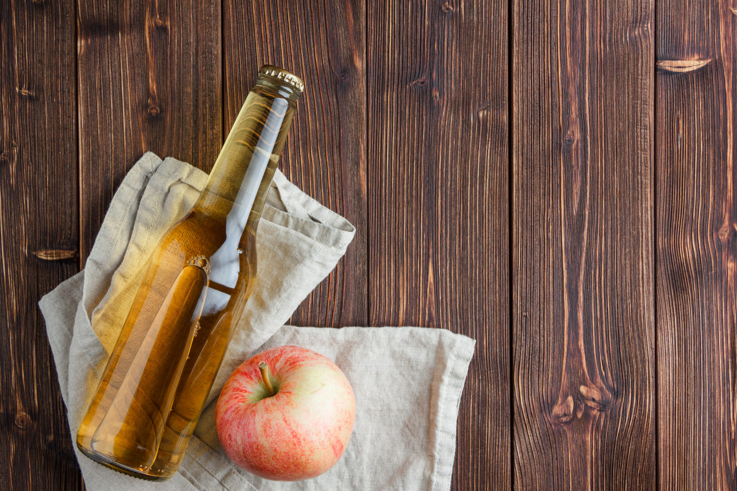 A bottle of hard cider rests on it's side next to an apple. The bottle and apple sit on a beige tea towel that's set out on a wooden background.