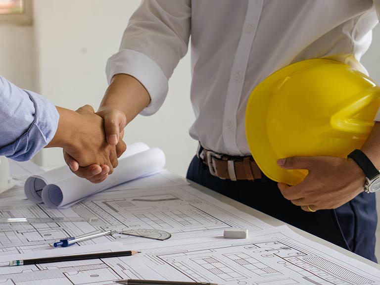 Two men shake hands over a table with blueprints. One man is holding a yellow hard hat under his arm.